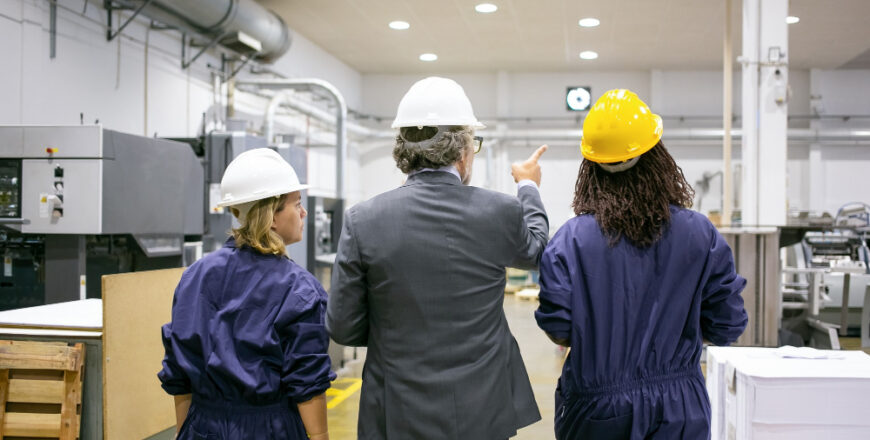 male-engineer-female-factory-employees-hardhats-walking-plant-floor-talking-man-pointing-equipment-instructing-women